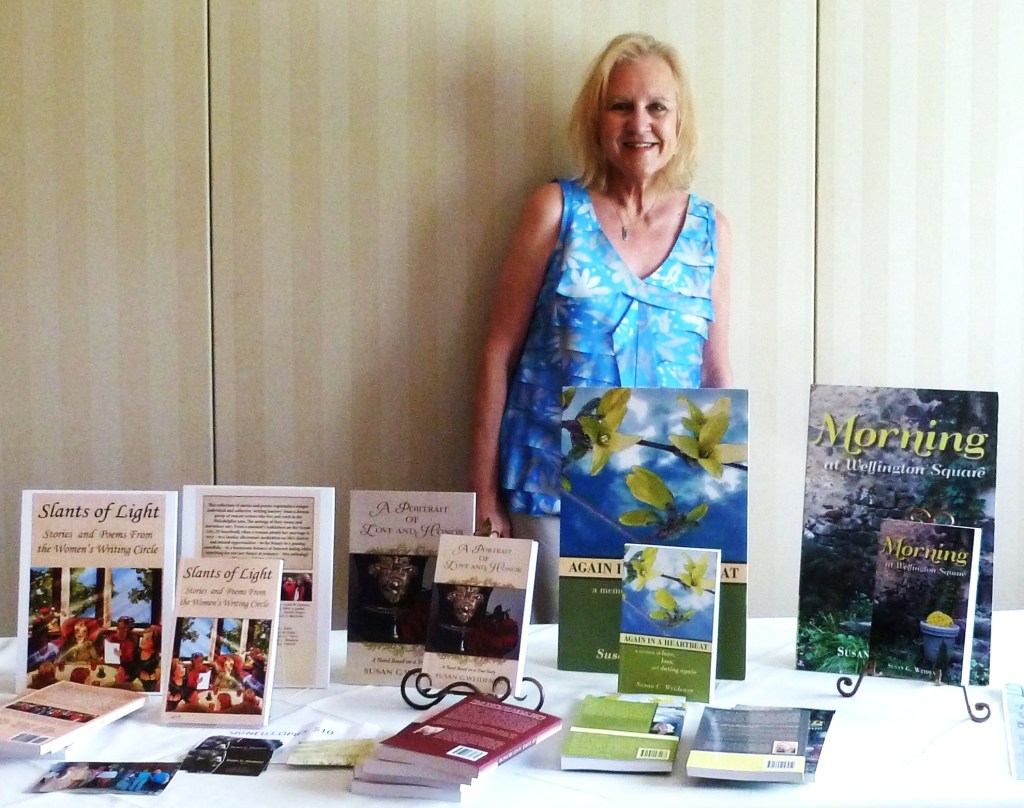 A woman in a blue floral top stands next to a display of books at a table, featuring titles like 'Slants of Light' and 'Morning at Wellington Square'.